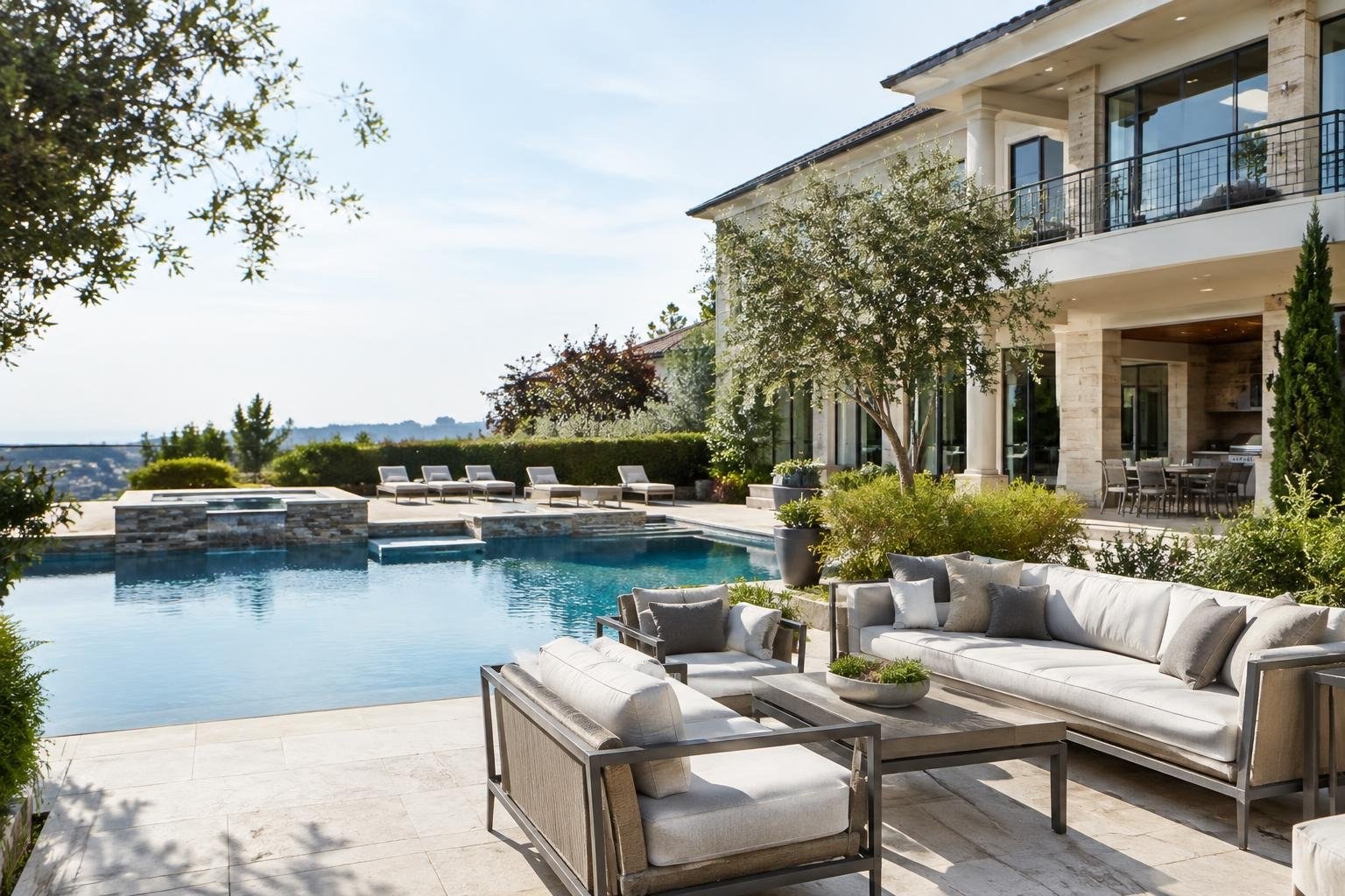 Homeowner and house manager reviewing a property operations dashboard A modern stone-and-glass house overlooks a swimming pool, with cushioned patio seating, lounge chairs, and a dining area among trees and shrubs.