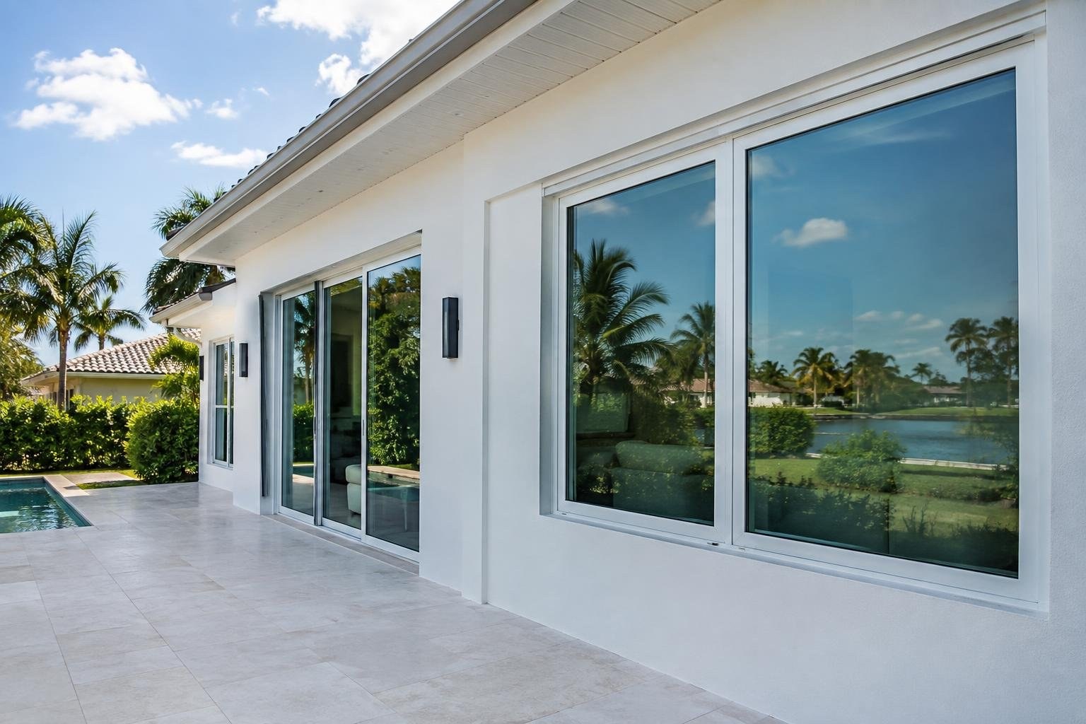 A white modern house with large reflective windows opens onto a tiled patio beside a swimming pool, with palm trees and a waterway visible around it.